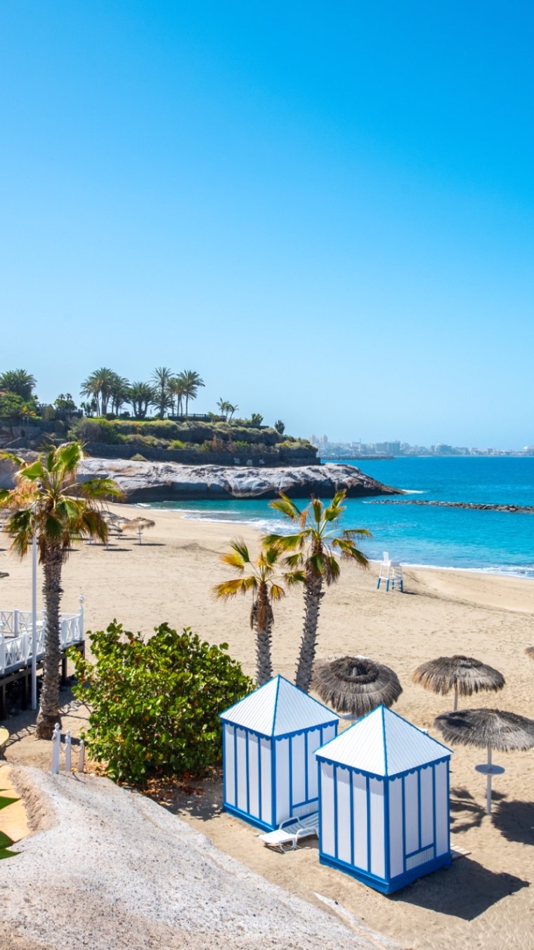 Playa del Duque en Tenerife: arena dorada, palmeras, casetas azules y blancas y mar turquesa